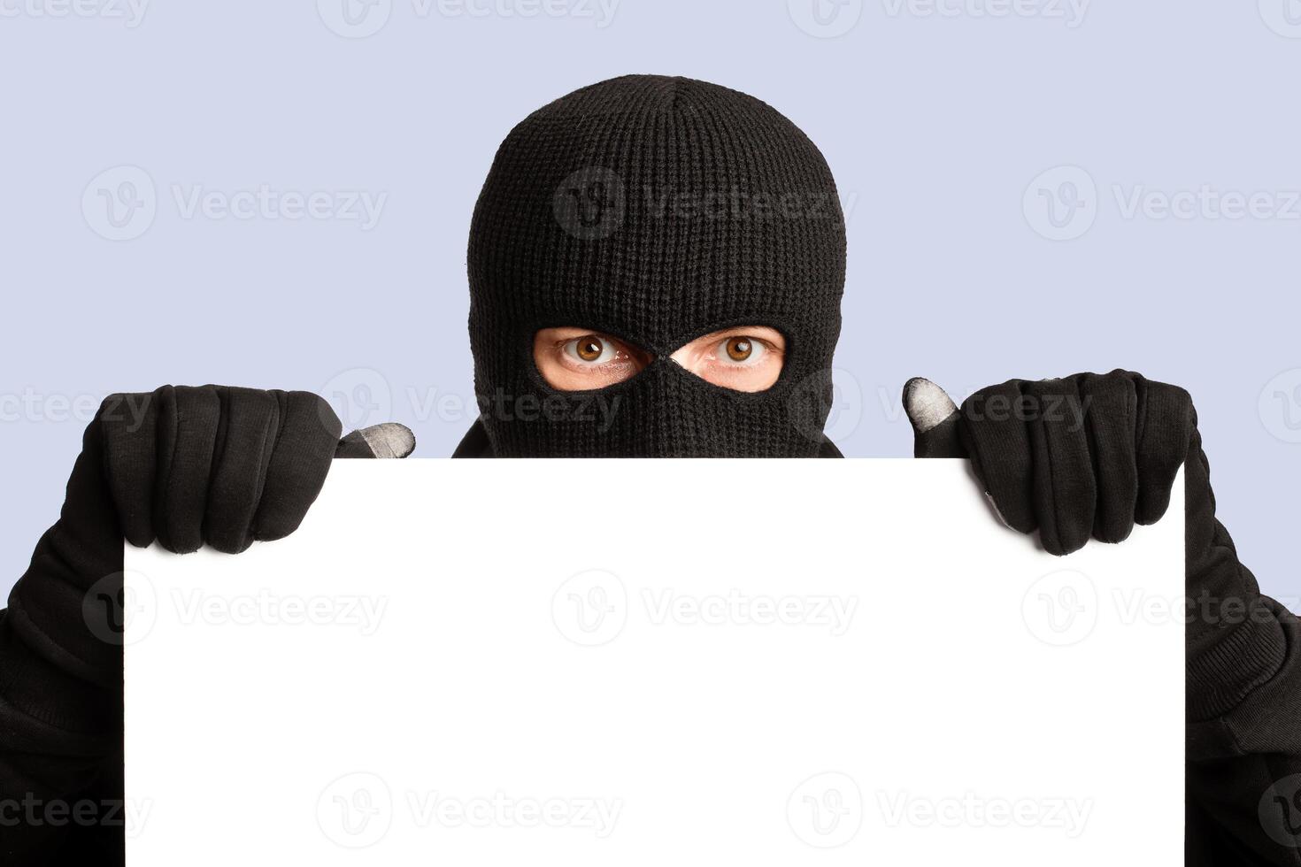 Masked gangster holding and looking out white blank board, isolated over white studio wall photo