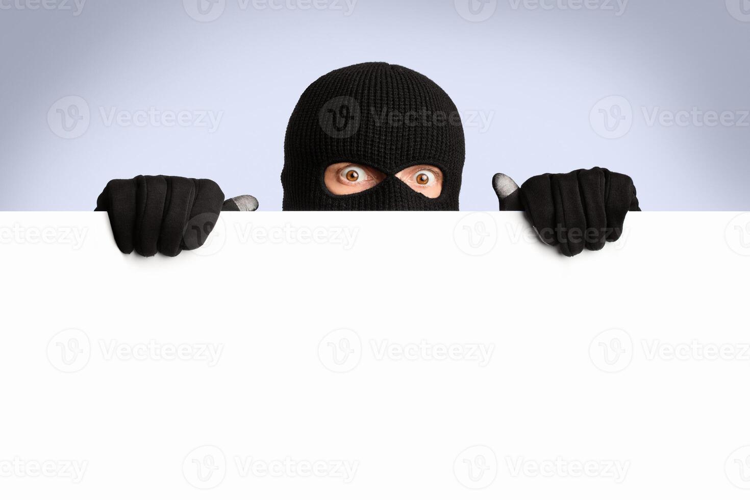 Scared and surprised masked gangster holding and looking out white blank board, isolated over white studio wall photo