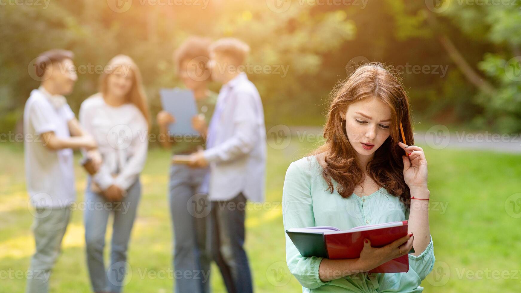 Focused redhead student girl preparing for lecture outdoors while her groupmates chatting on background, selective focus photo