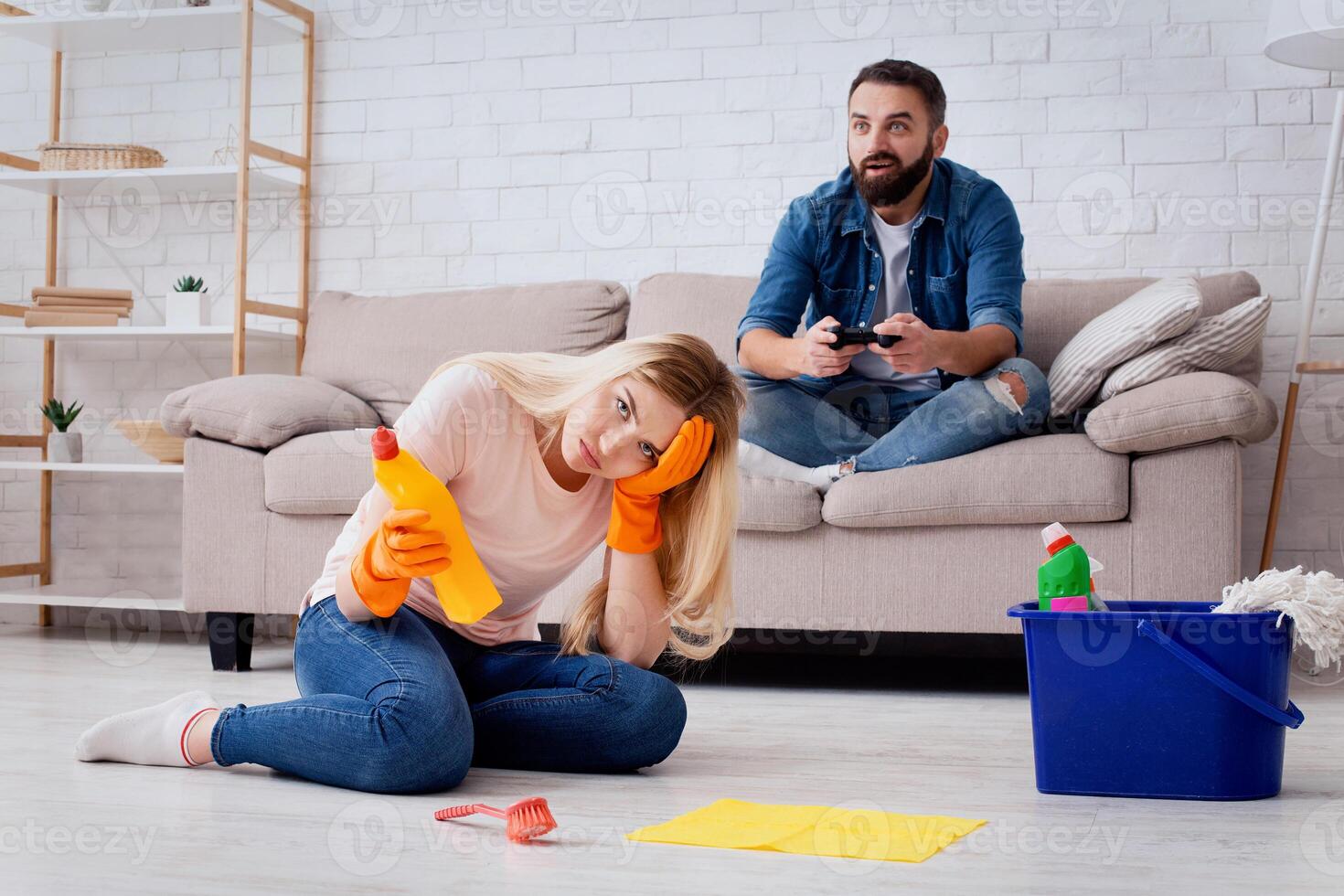 A woman sits on the floor looking stressed while cleaning, while a man sits on the couch playing games. They are in their living room, highlighting tensions in their marriage. photo