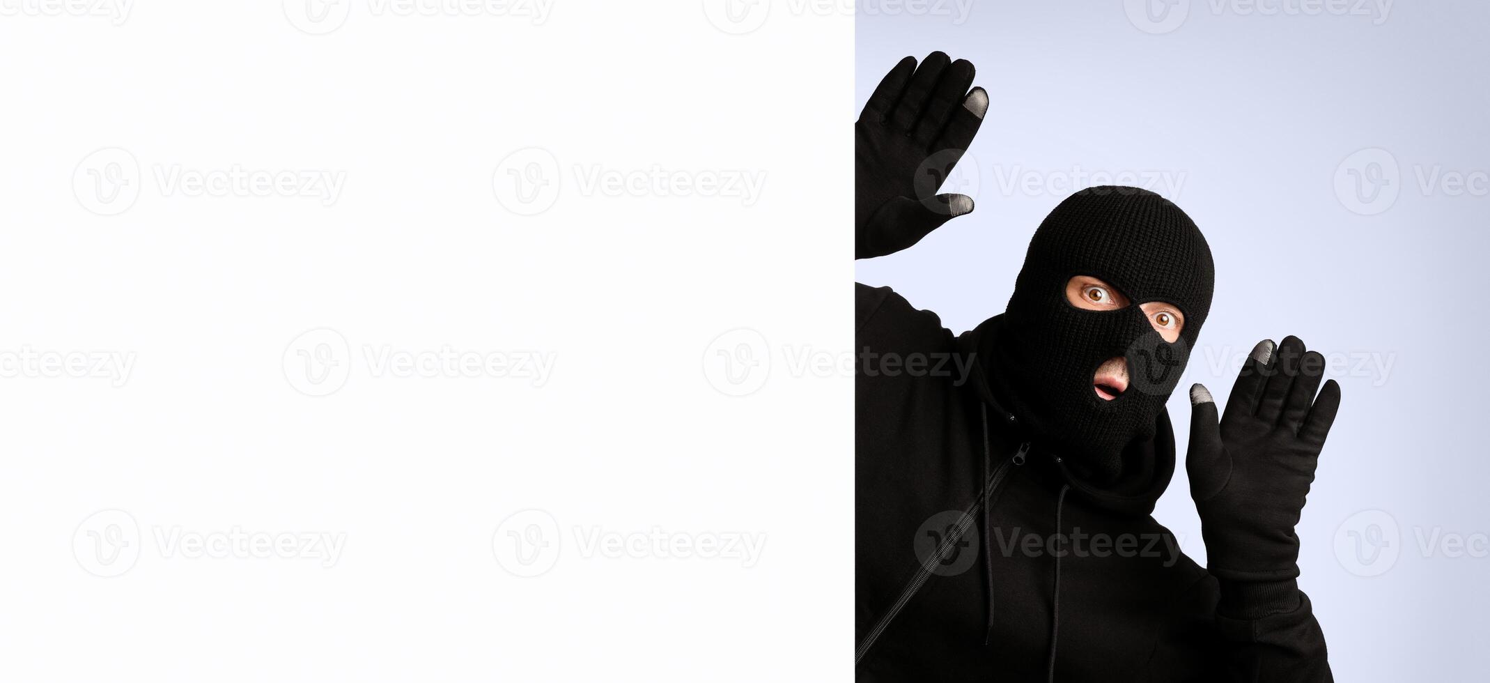 Scared thief in a balaclava and gloves raising hands up and looking out of blank sign with copyspace for your text photo