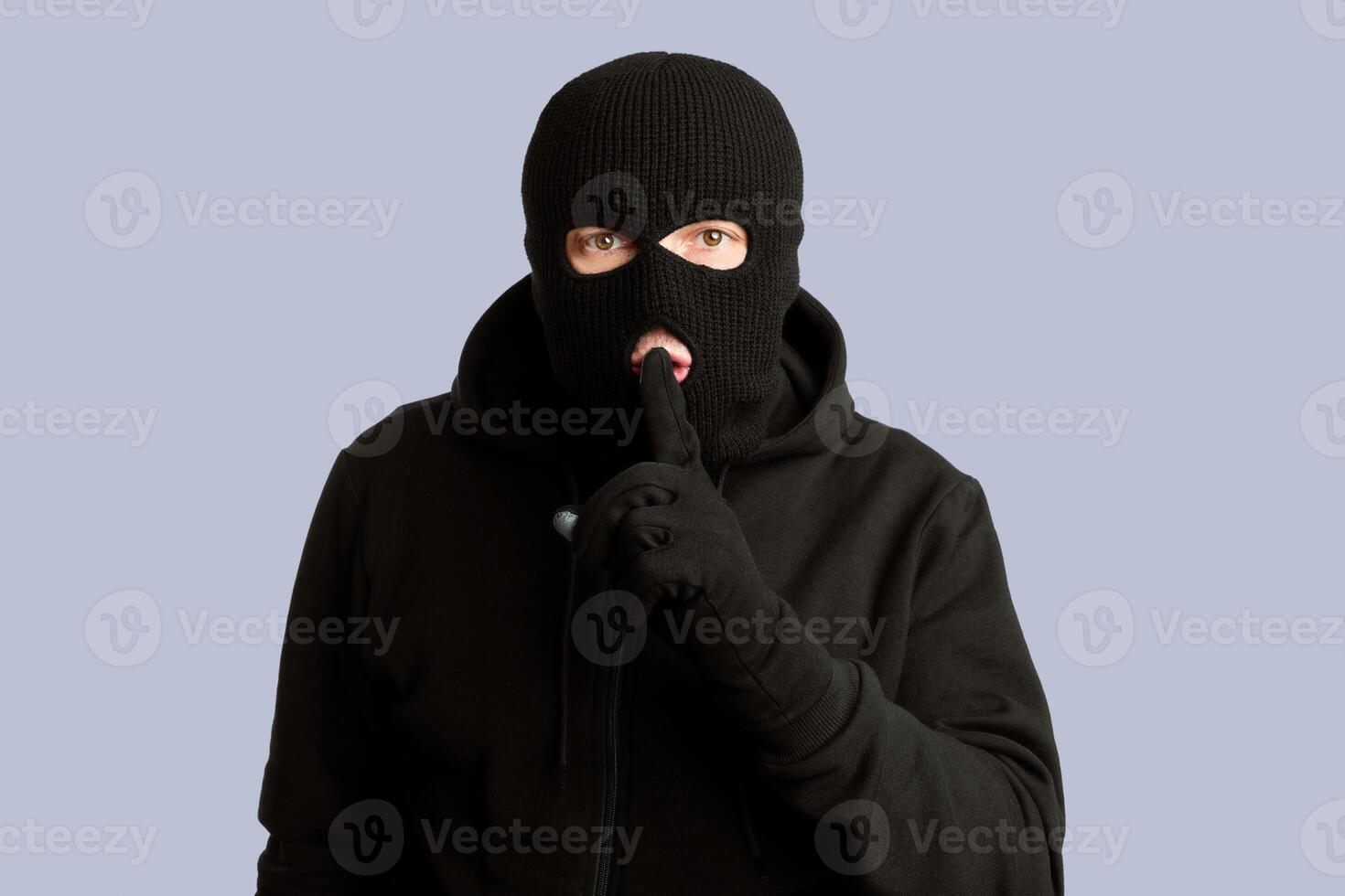 Keep Silence Concept. Portrait of thief in black balaclava holding fingers on lips, making hush sign, isolated on white wall photo