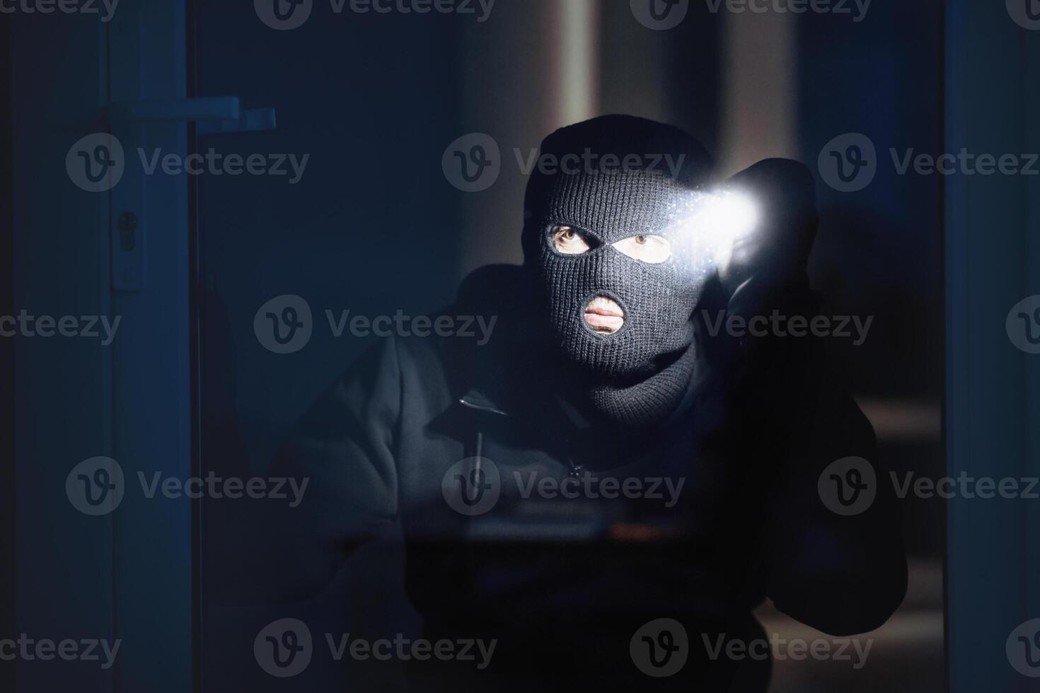 Spying Concept. Burglar in black balaclava hat watching what's inside the building in the night, using torch photo