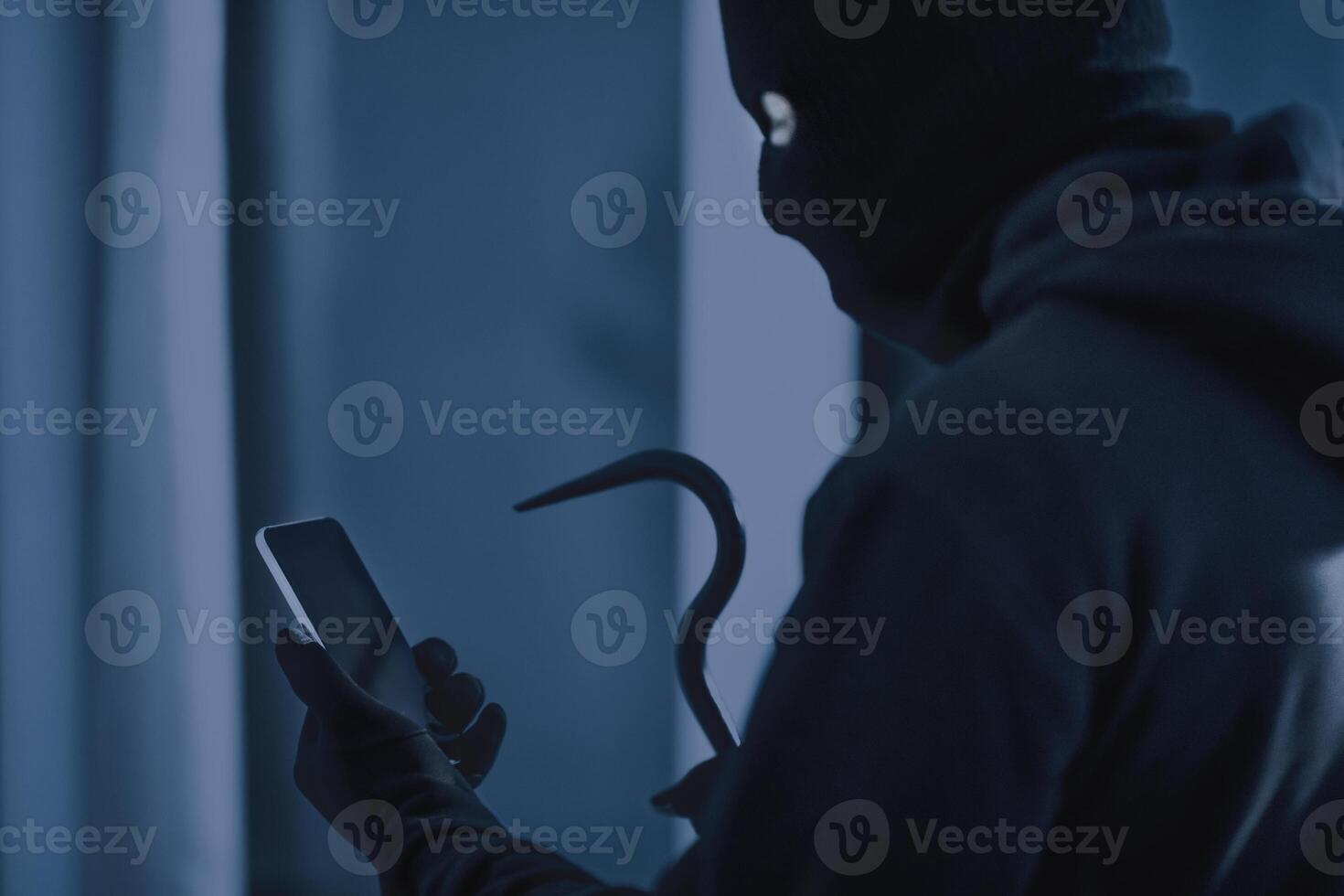 Robbery Concept. Masked villain with crowbar and smartphone preparing burglary via smart home app in the dark photo