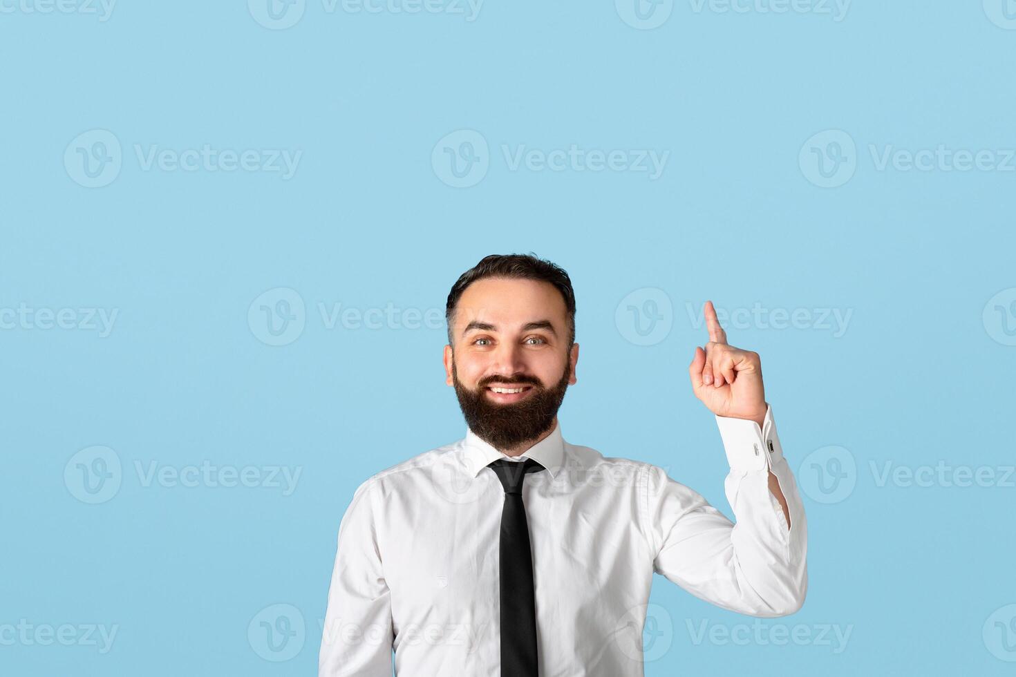 Happy millennial businessman in formalwear pointing upwards at empty space on blue studio background. Attractive bearded CEO having creative idea, gesturing eureka, experiencing aha moment photo