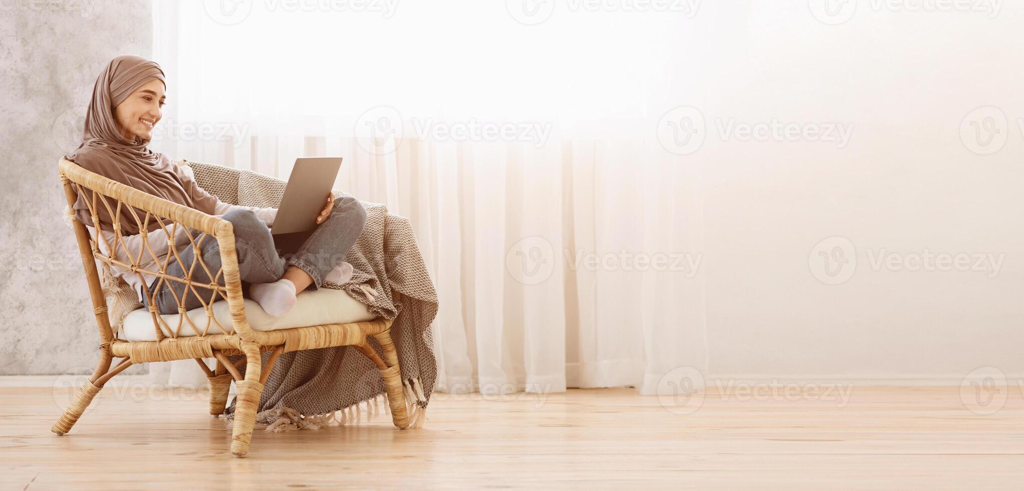 Weekend Leisure. Happy muslim girl in hijab using laptop while relaxing in wicker chair at home, working remotely or watching movies online, panorama photo