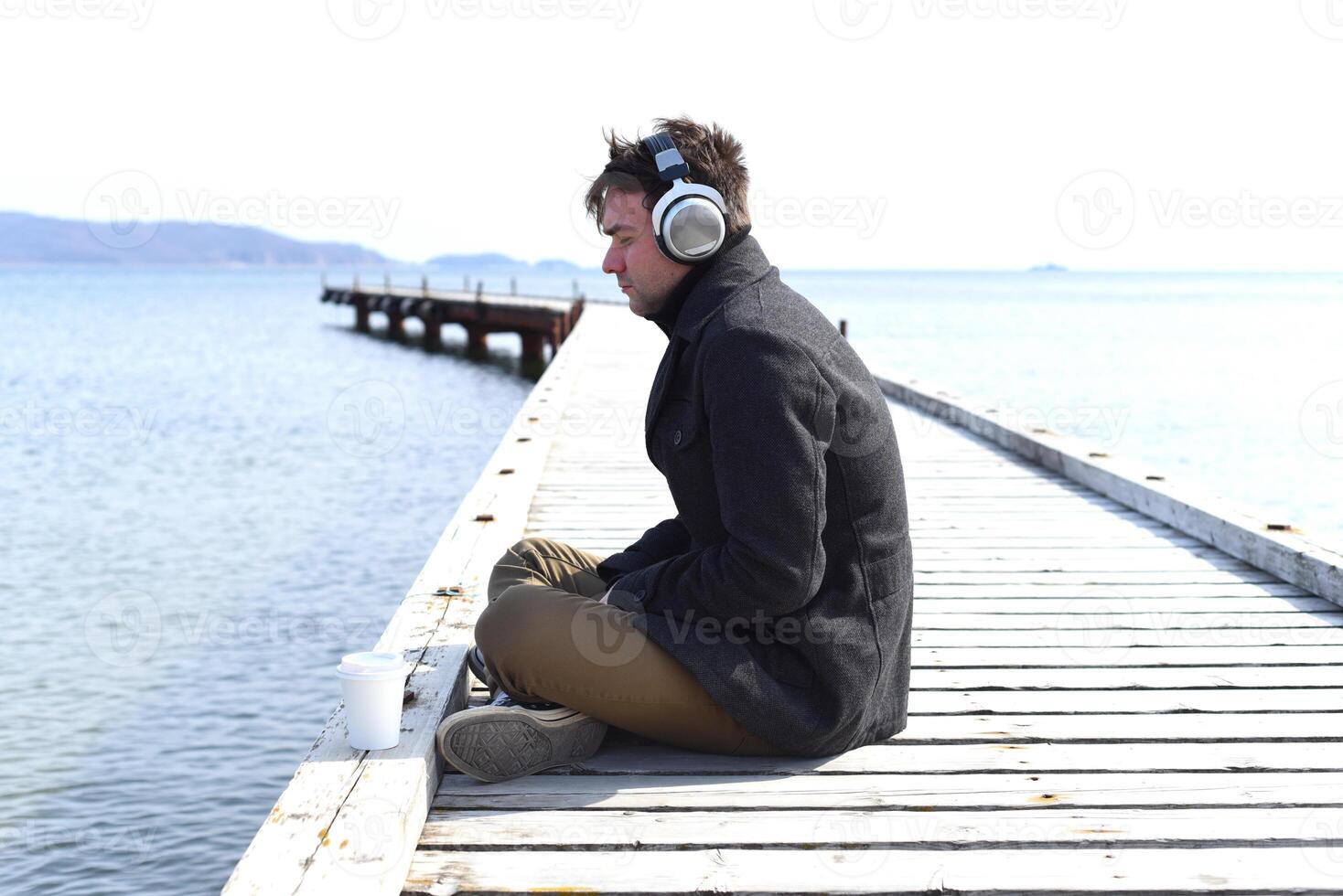 Young gloomy man listening to music sitting all alone on pier on a windy spring day. photo