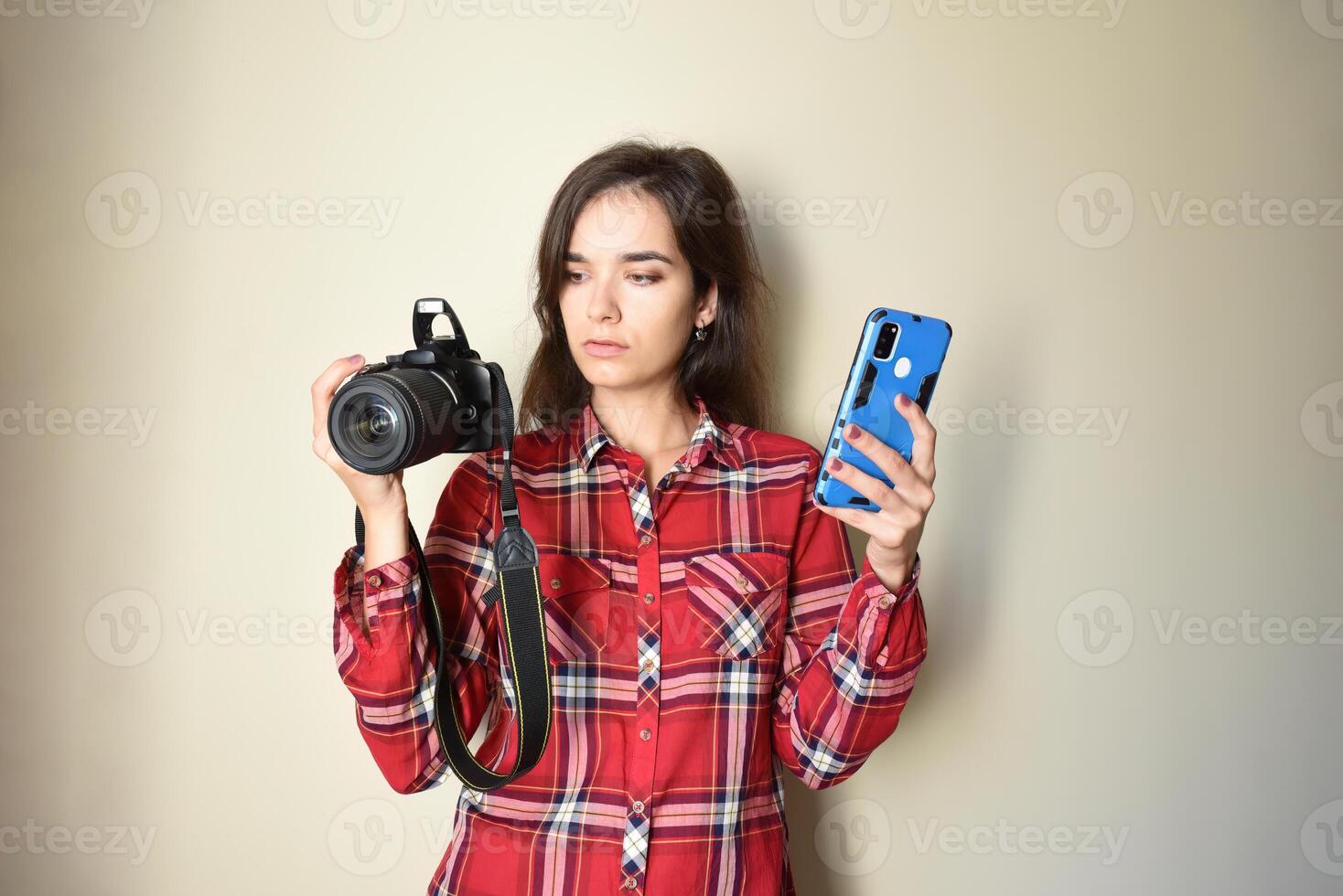 Young brunette woman photographer in red shirt choosing between camera and mobile phone on beige background. Concept of choice and difference between mobile photography and camera. Copy space photo