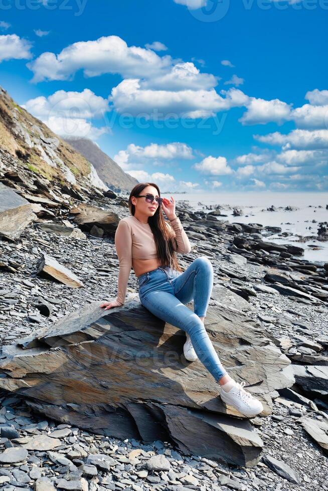 Young russian brunette woman in beige blouse sitting ont the stone near Japanese sea on a sunny day photo