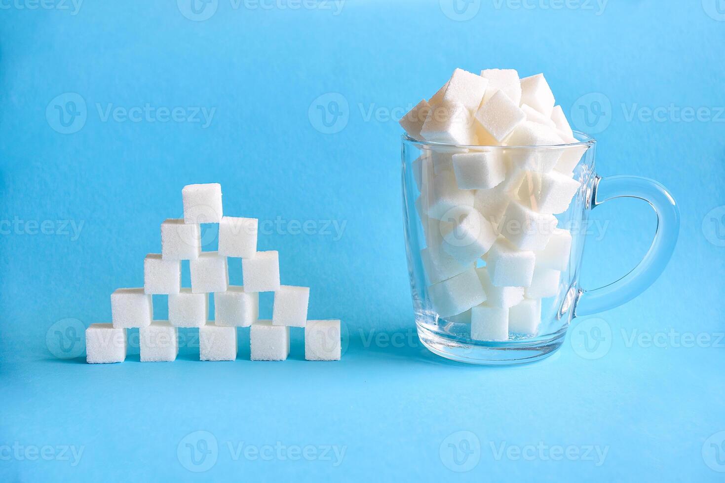 Transparent mug full of refined white sugar cubes and pyramid made from cubes of white sugar on blue background. Copy space photo