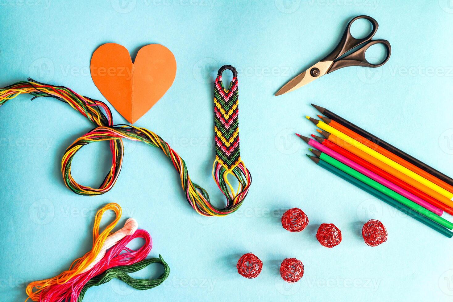 Unfinished woven friendship bracelet with skein of thread, scissors, cut heart and selected palette of crayons colors on white background photo