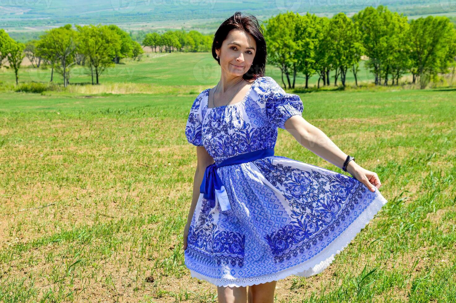 Slender 45 year old Russian smiling brunette woman in blue dress in russian slavic style on a green field. Copy space photo