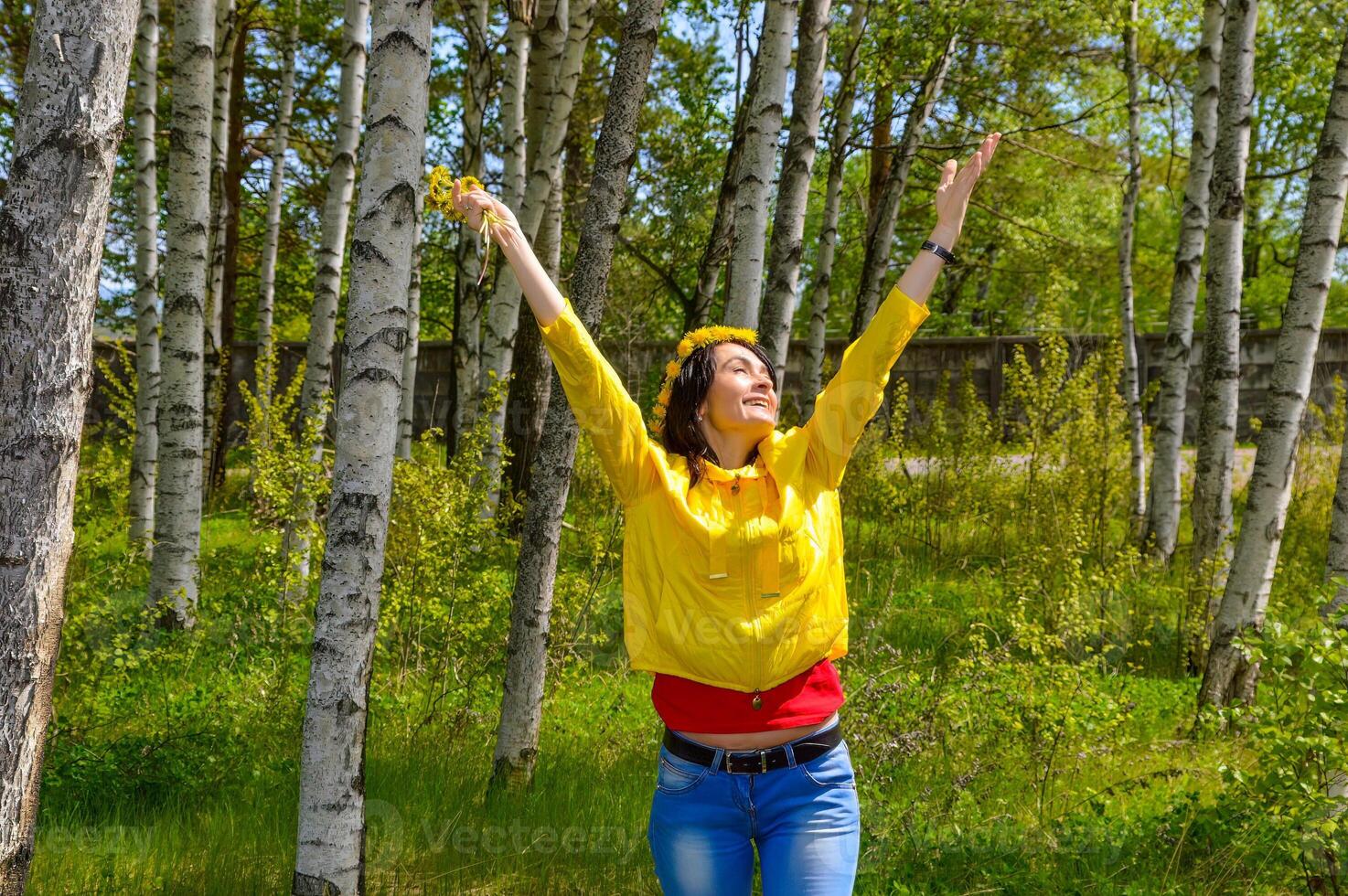 Brunette woman with bouquet of dandelion and wreath embracing nature with open arms in countryside. copy space photo