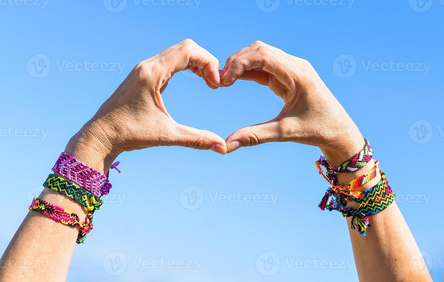 Hands making heart symbol on sky background. friendship bracelets on wrists photo
