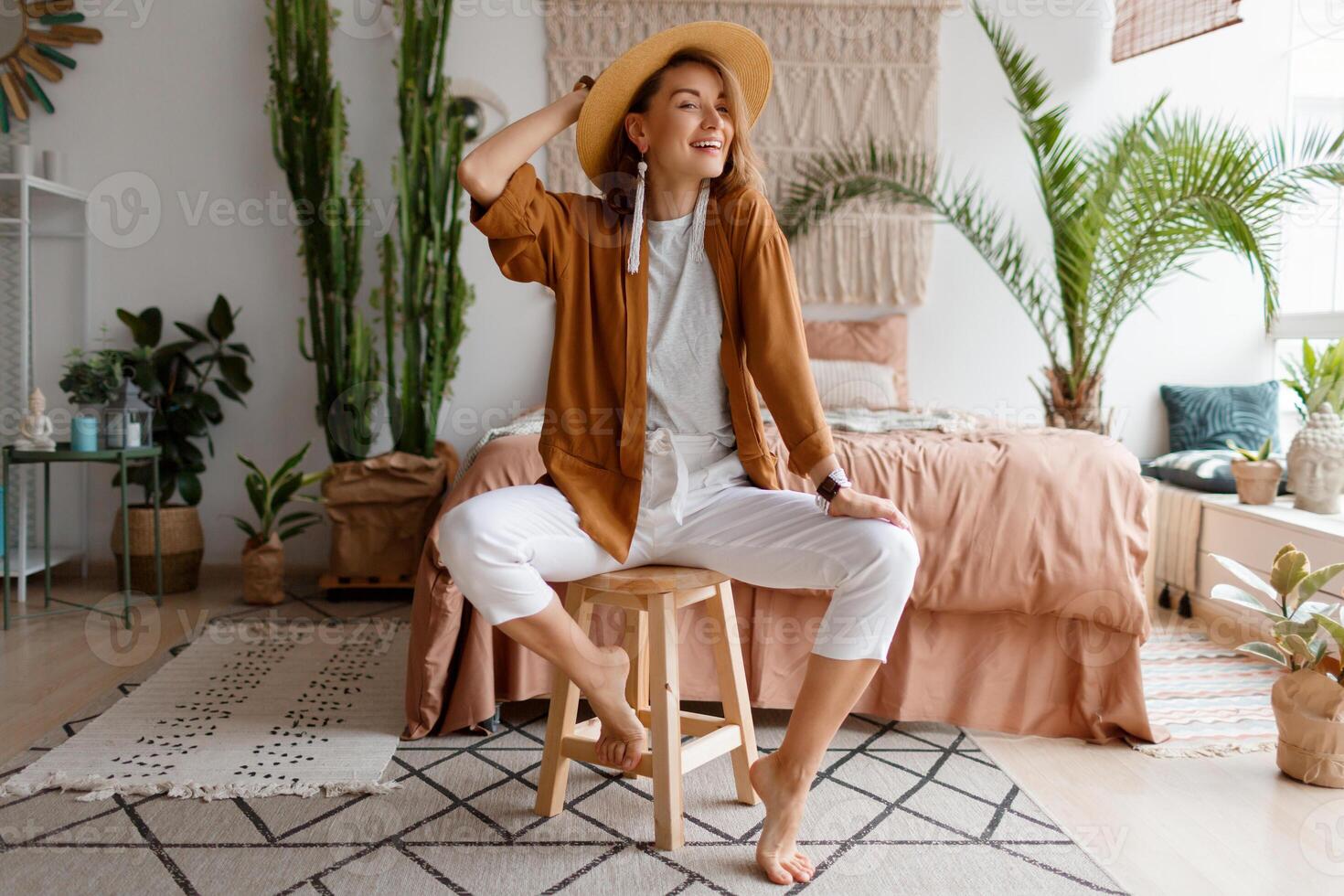 Romantic woman with candid smile sitting on bed, enjoying sunny morning in her stylish flat in boho style. photo
