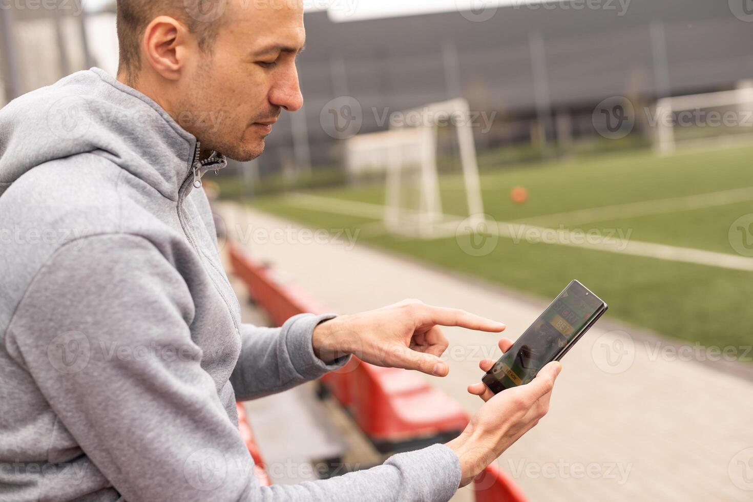 Portrait of handsome smiling bearded man being happy to win at bookmaker's mobile application after betting at favourite football team, stadium on the background photo