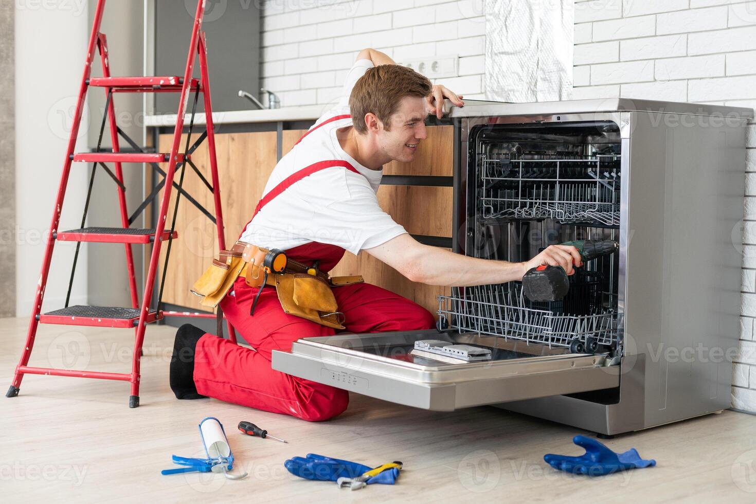 Technician or worker in uniform installs dishwasher into the kitchen furniture. Repairman wear worker suit repairing maintenance of dishwasher. Master in protective gloves fix dishwasher. photo