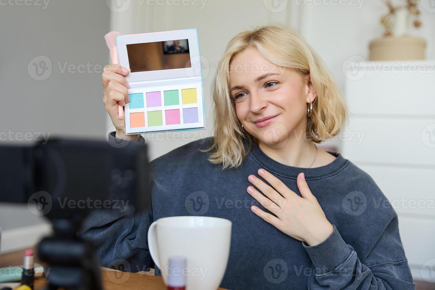 Lifestyle blogger, records in her room, has a camera on coffee table, shows eyeshadow palette to her followers, does makeup tutorial, vlogger working indoors, creates content for social media photo