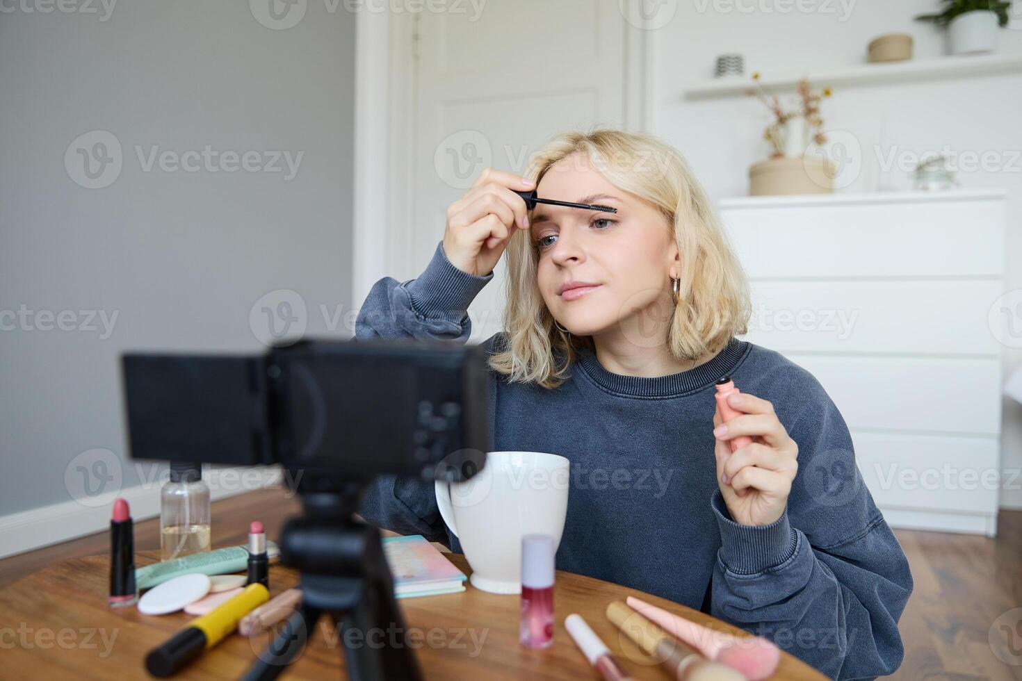 Portrait of beautiful, smiling blond woman, girl recording of her makeup tutorial for social media, vlogger sitting on floor in her room, using stabiliser to create content, reviewing mascara photo