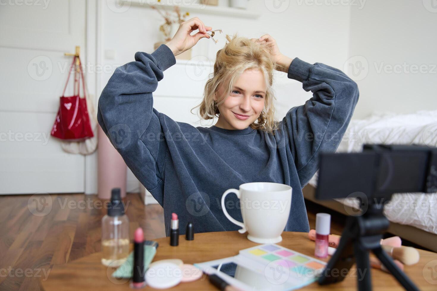 Lifestyle blogger records of herself while doing hairstyle, showing makeup lifehacks for social media followers, vlogging, using camera for her blog, sitting in a room, drinking tea photo