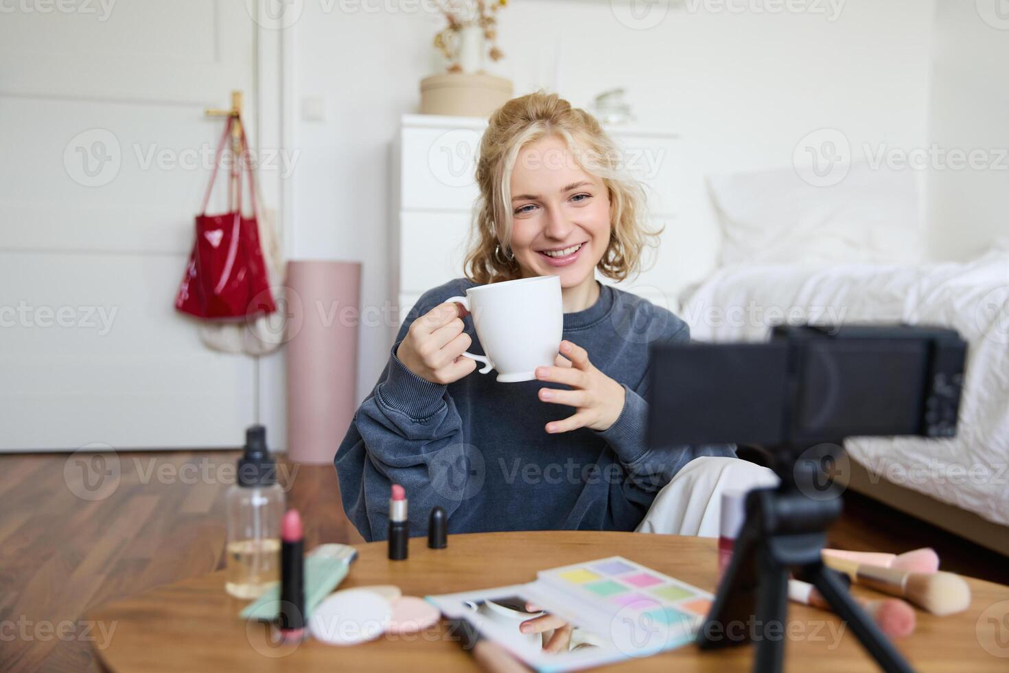 Cheerful woman, beauty blogger, records lifestyle vlog on digital camera, talks casually, tells a story for social media followers, holds cup, drinks tea and sits on floor in her room photo