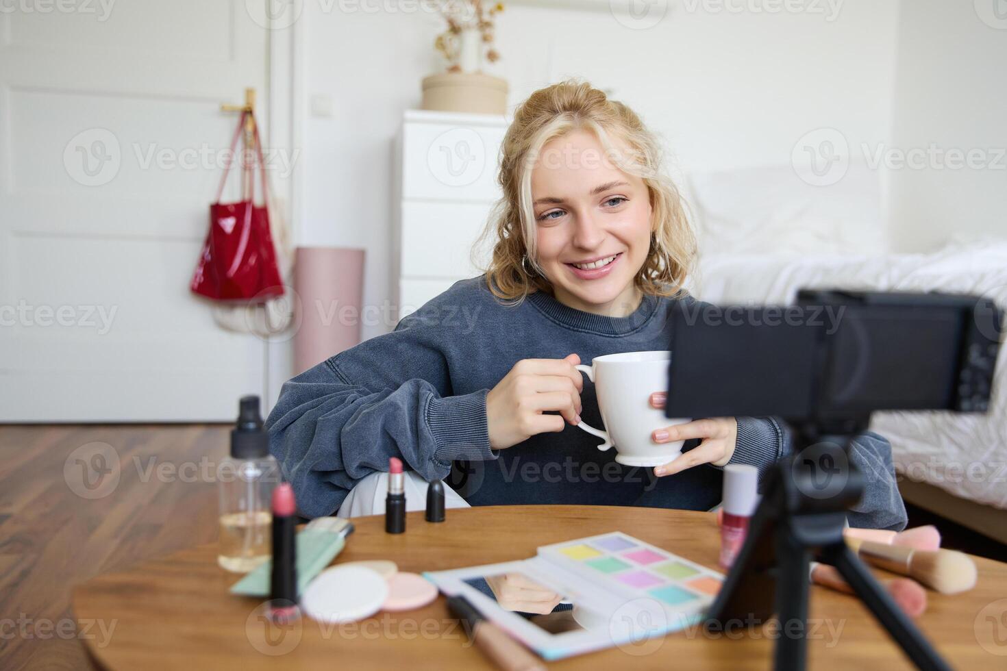 Blogging, modern technology, e-commerce and people concept. Happy smiling woman beauty blogger holding cup of tea applying makeup while recording beauty tutorial at home for her blog photo