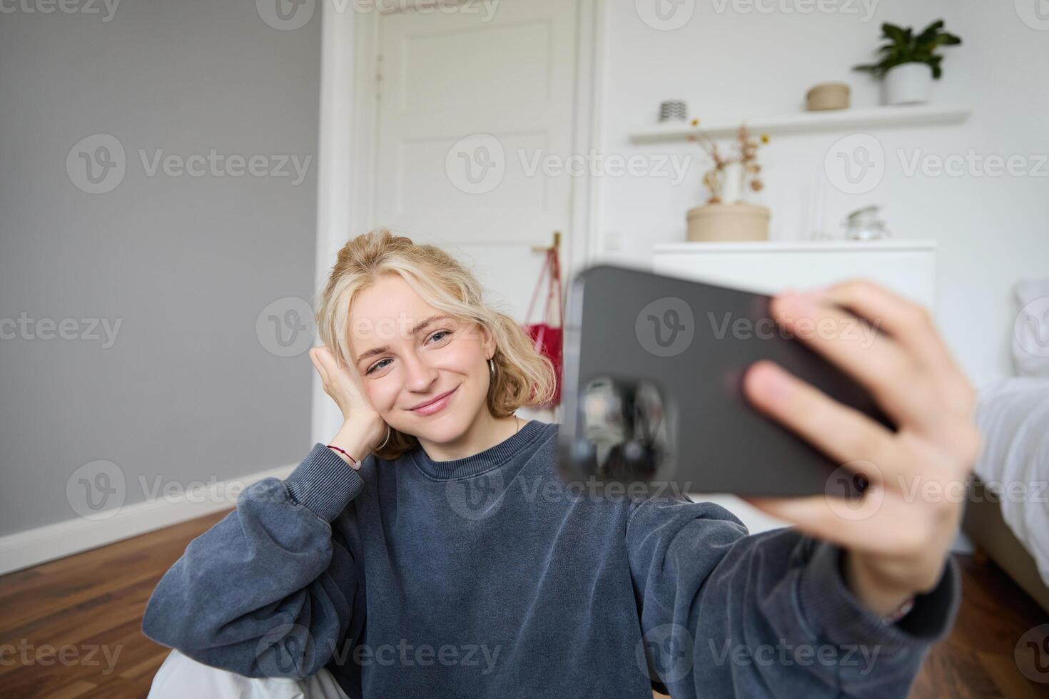 Portrait of young stylish girl sits on bedroom floor, takes selfies on her smartphone, posing for photo on social media app, smiling and looking happy at camera