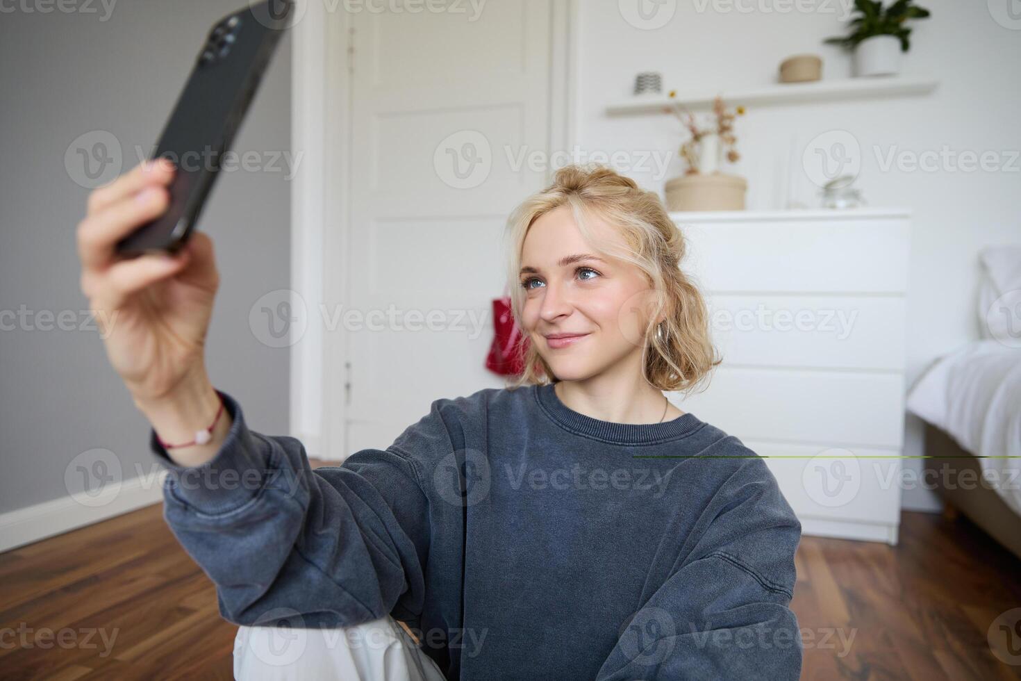 Portrait of young stylish girl sits on bedroom floor, takes selfies on her smartphone, posing for photo on social media app, smiling and looking happy at camera