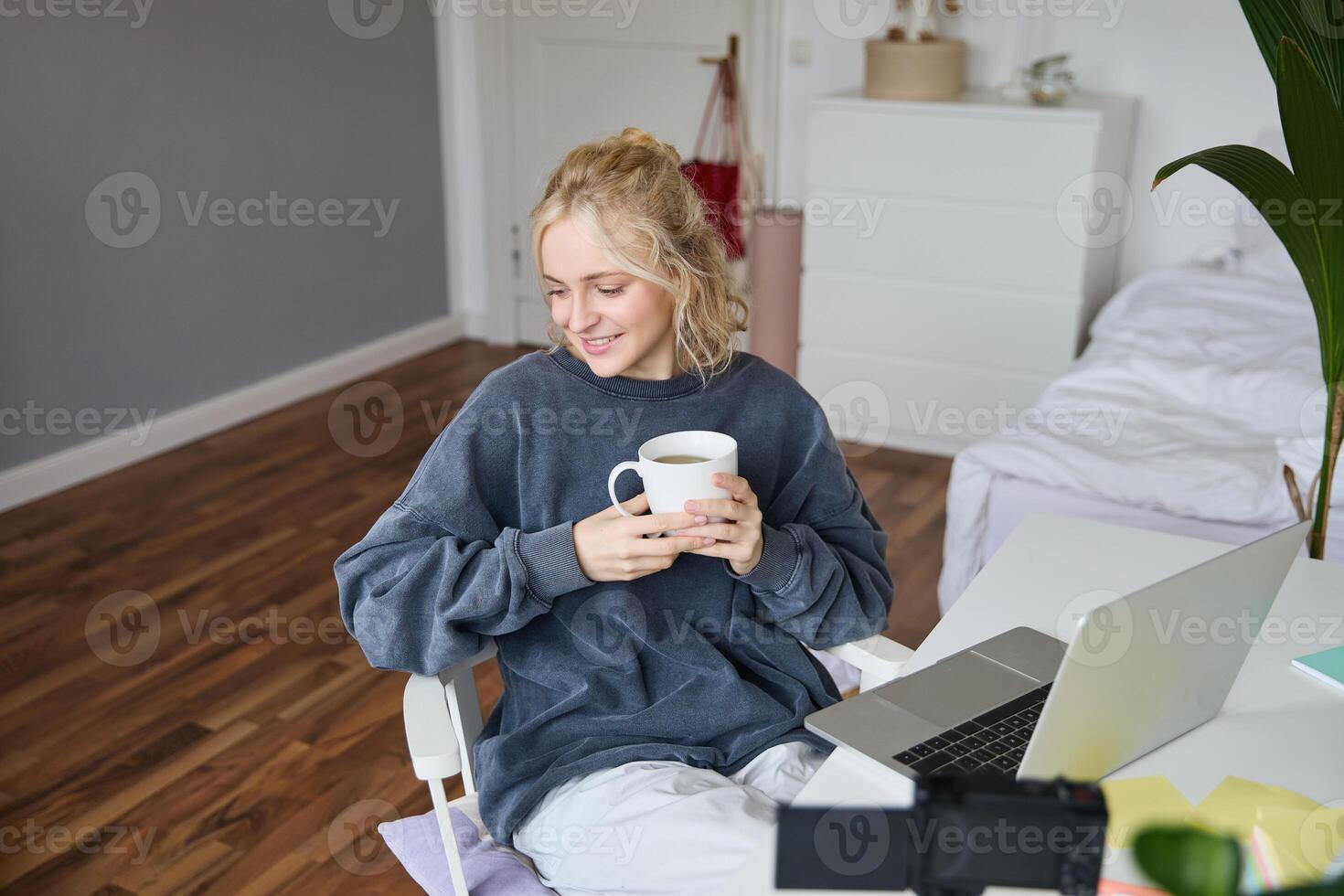 Portrait of young smiling woman, vlogger recording lifestyle in her room, sits in front of laptop and digital camera, drinks tea, creates content for social media account photo