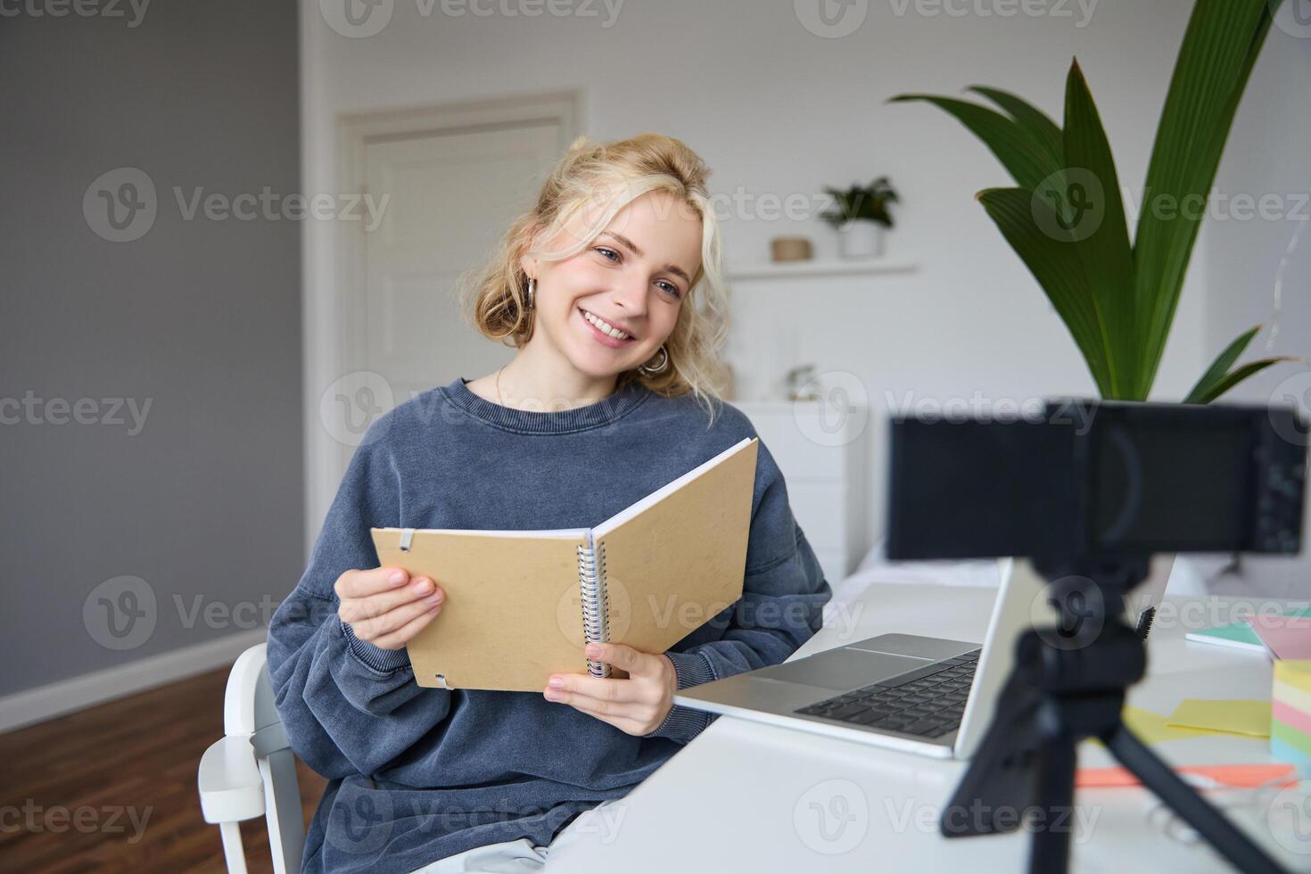 Portrait of cute young woman, teenage girl records on digital camera, uses laptop to create lifestyle content, shows notebook, reads notes, using computer in her room photo