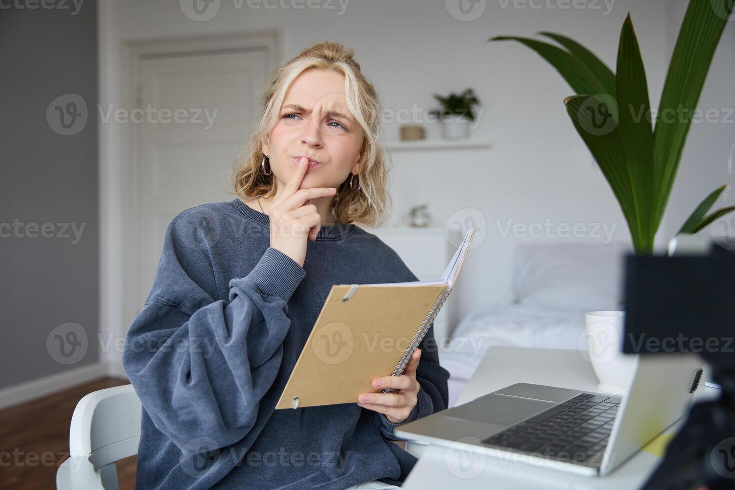 Portrait of young woman, student sitting in room, studying from home, remote education concept, holding notebook and thinking, looking aside with thoughtful face, concentrating on assignment photo