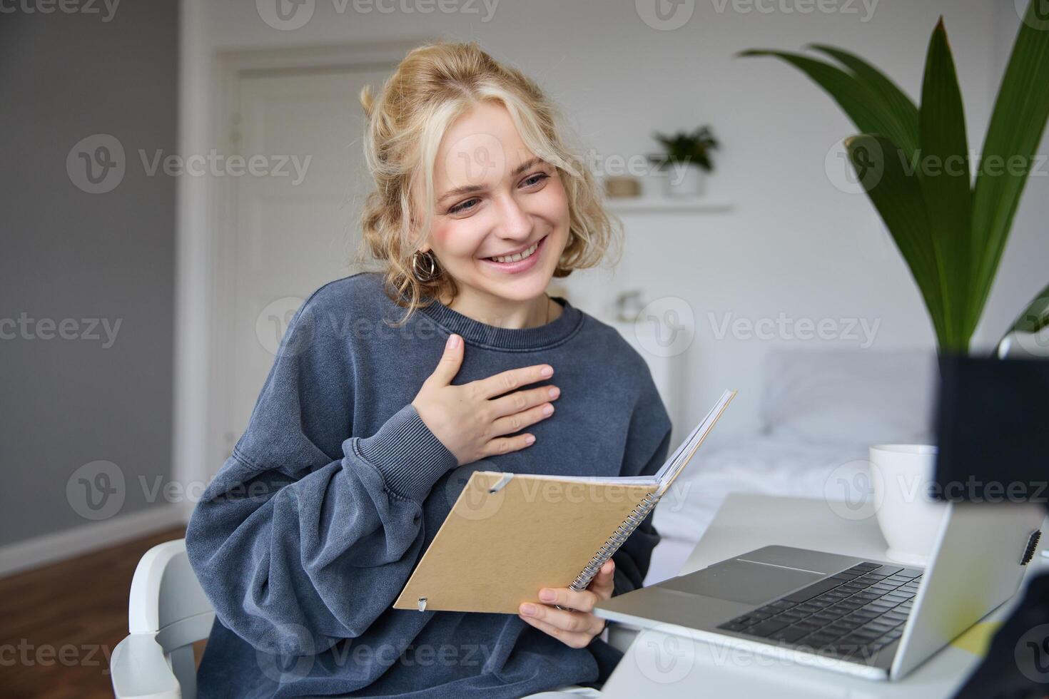 Portrait of smiling, beautiful young blond woman, student working on assignment from home, online learning in her bedroom, talking to camera, chatting, holding notebook photo