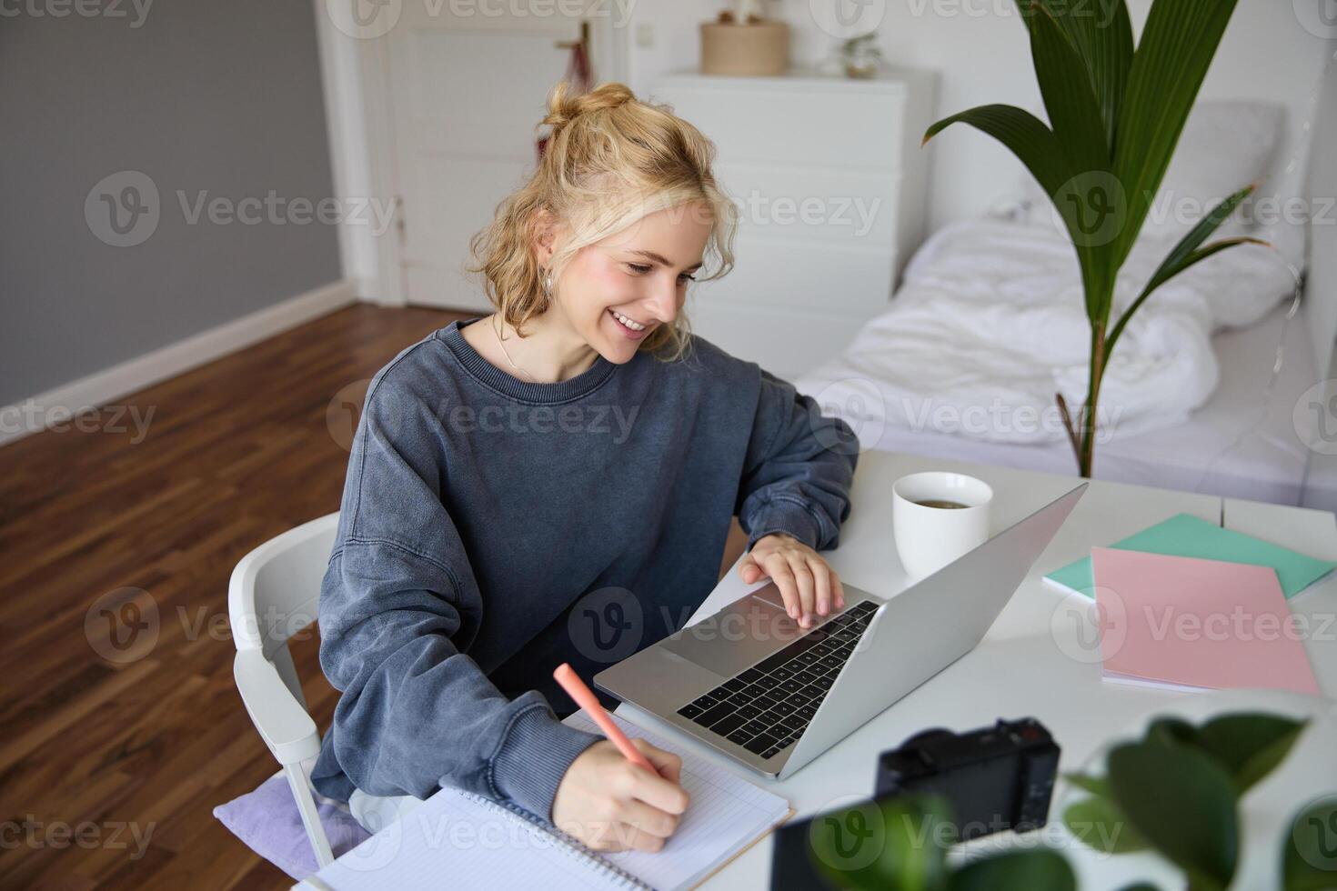 Portrait of smiling young woman, female student, doing distance learning course, using laptop, studying at home, writing down, making notes photo