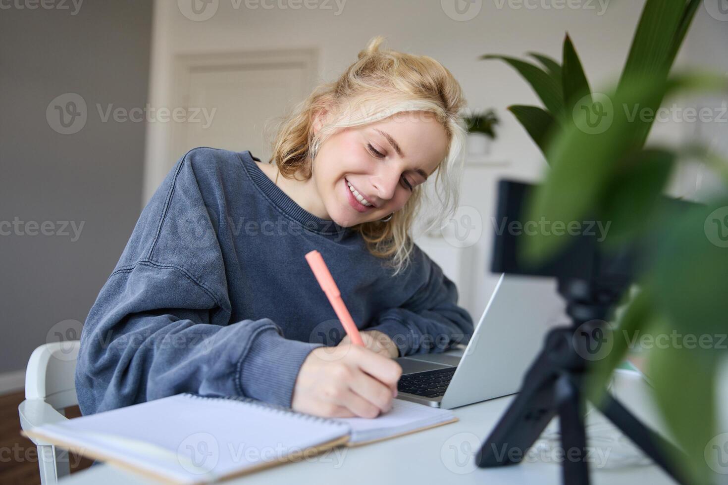 Portrait of young woman, lifestyle blogger, recording of herself, making notes, writing in journal, sitting in front of laptop in a room and studying photo