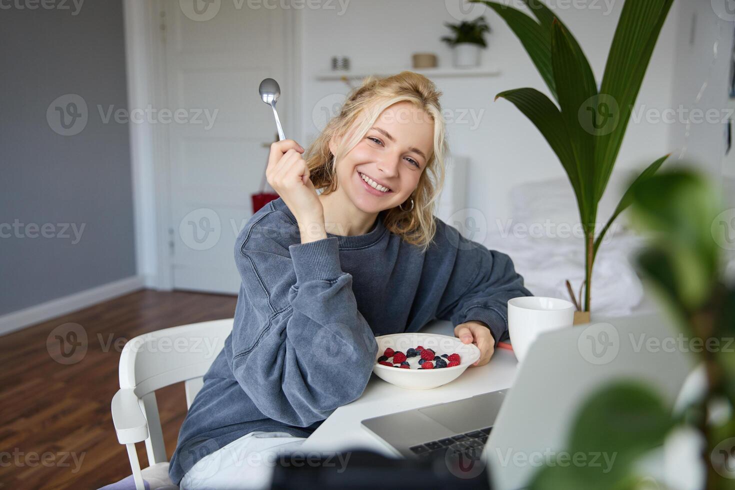 Portrait of smiling blond young woman, eating in front of laptop, watching s online while having breakfast, enjoying dessert, sitting in bedroom photo