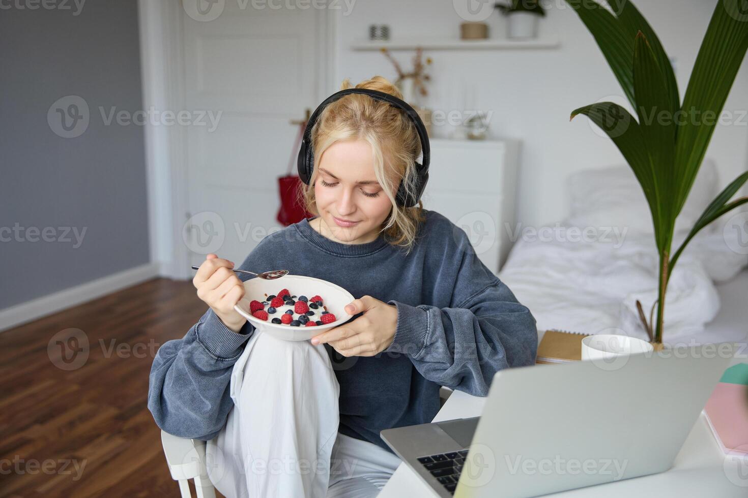 Portrait of smiling young blond woman in headphones, sitting in room, watching movie on laptop, eating breakfast and drinking tea, having lunch in front of computer screen photo