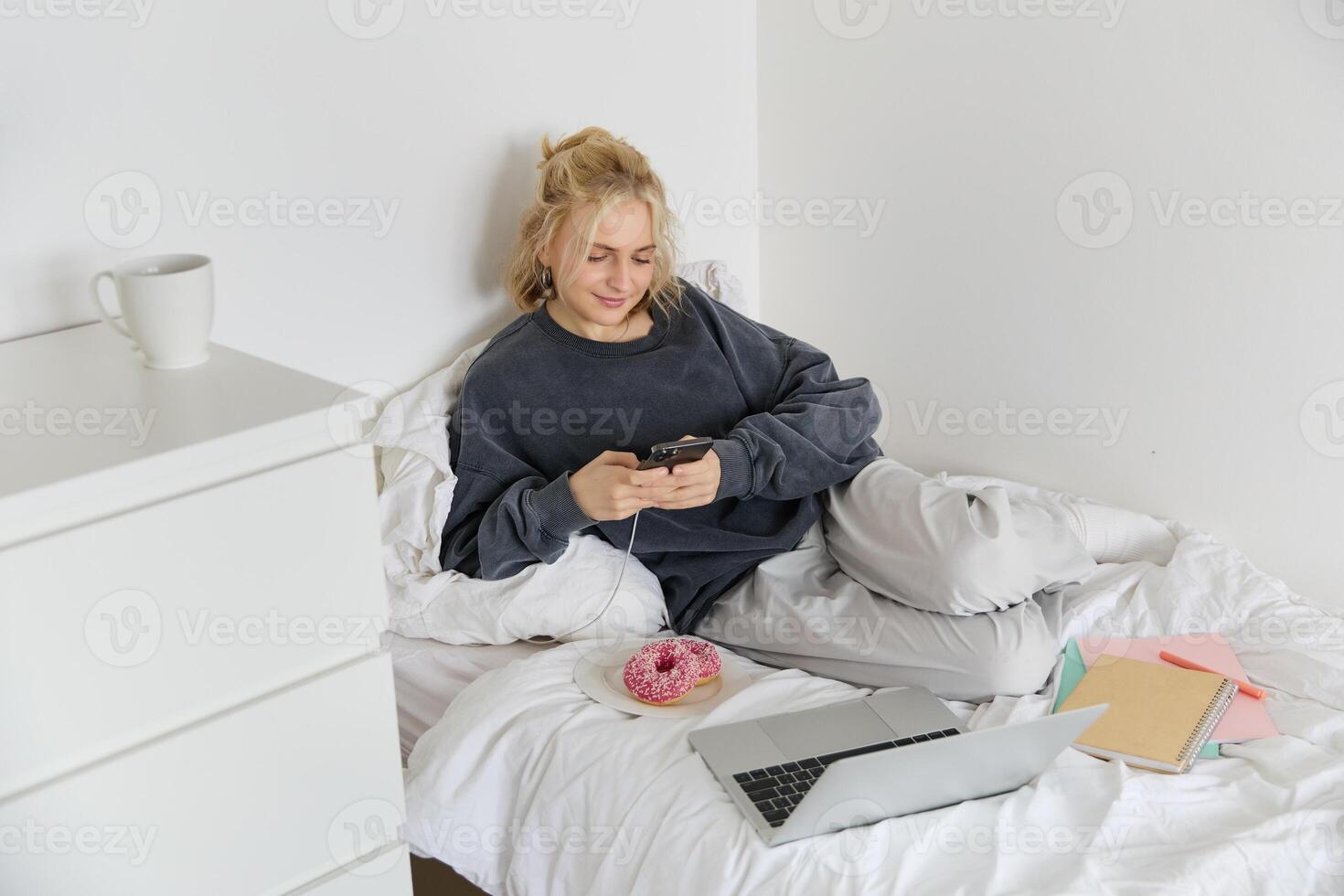 Portrait of smiling candid woman, lying in bed with doughnut, using smartphone and laptop, resting at home in bedroom, watching tv show or chatting online photo