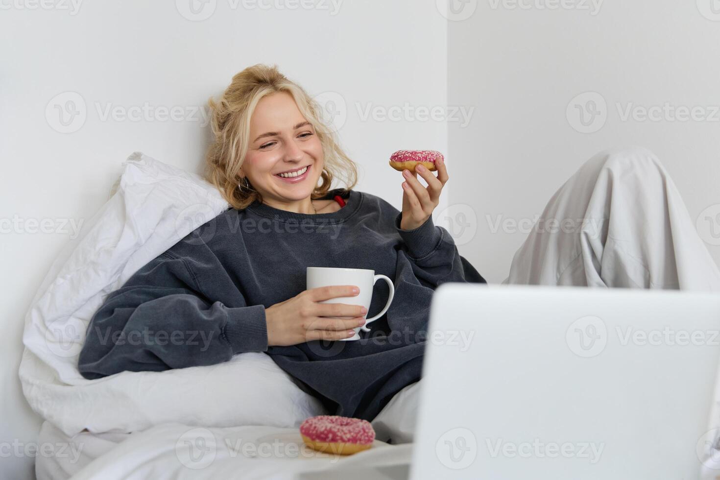 Lifestyle and people concept. Happy blond woman lying in bed with food, drinking tea and eating doughnut, resting at home, spending time alone, watching tv show photo