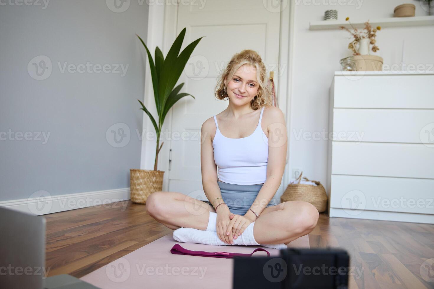 Portrait of young fitness instructor, vlogger showing exercises on camera, recording herself, sitting on mat with laptop, doing workout, explaining yoga movements to followers photo