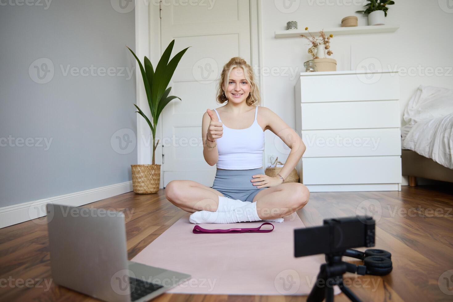 Fitness blogger, woman records of herself, sitting on rubber mat for exercises in front of digital camera and laptop, shows thumbs up, creates content for social media account about wellbeing photo