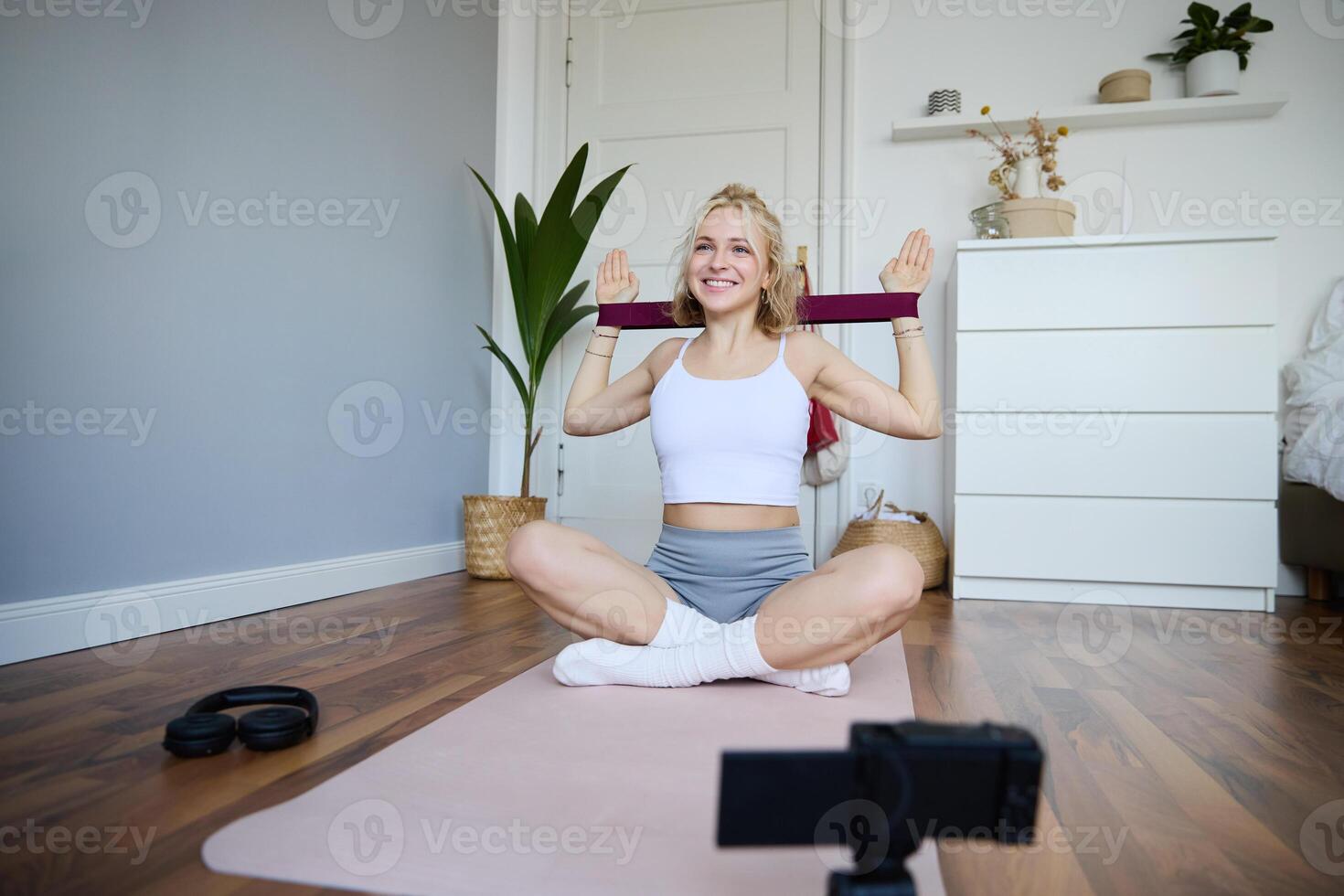Portrait of young blond woman, social media vlogger using digital camera during workout, shooting about exercises and fitness, stretching with rubber resistance band photo