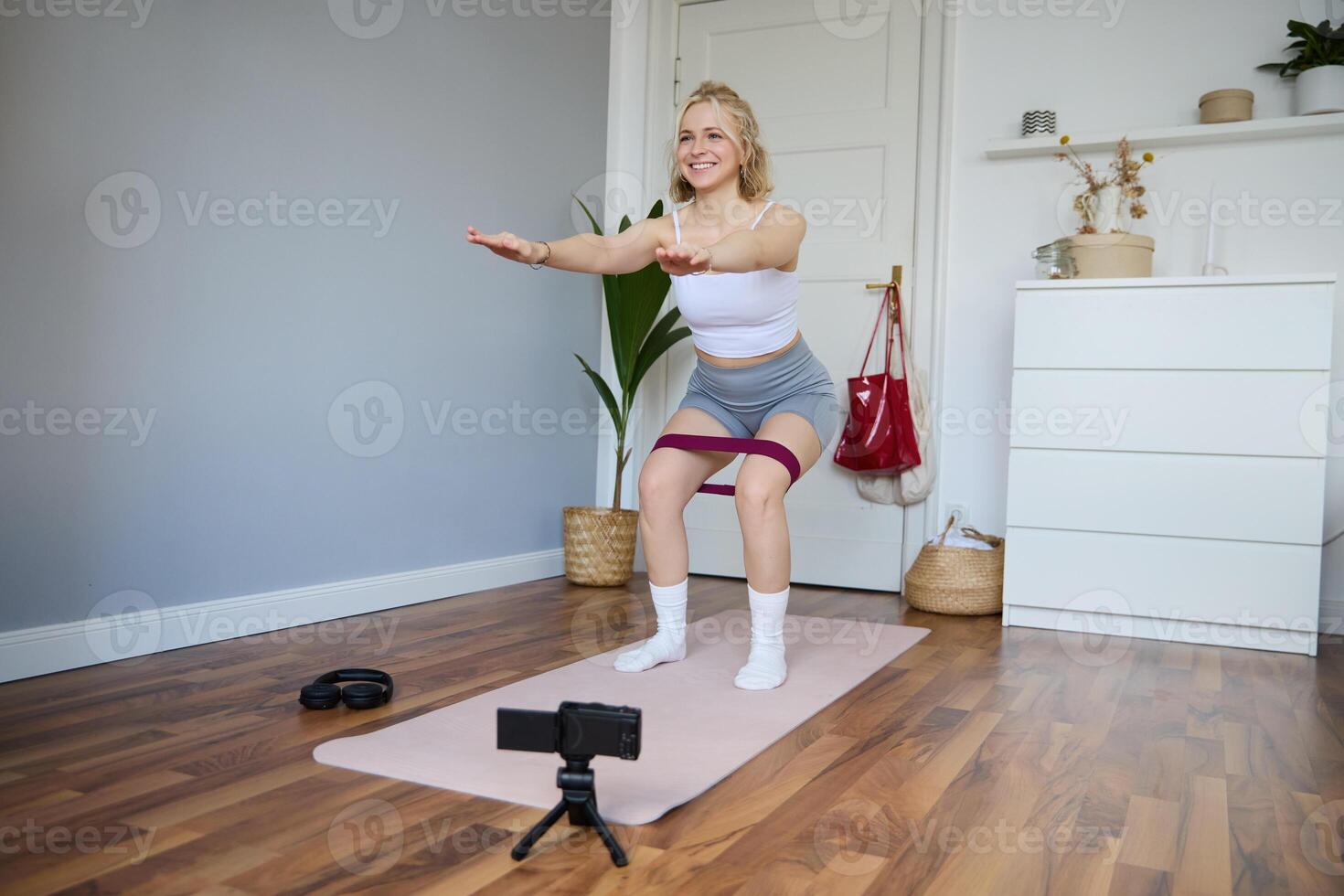 Portrait of woman, fitness instructor at home, recording about workout, showing how to do leg exercises, squats with elastic resistance band, working out indoors on yoga mat photo