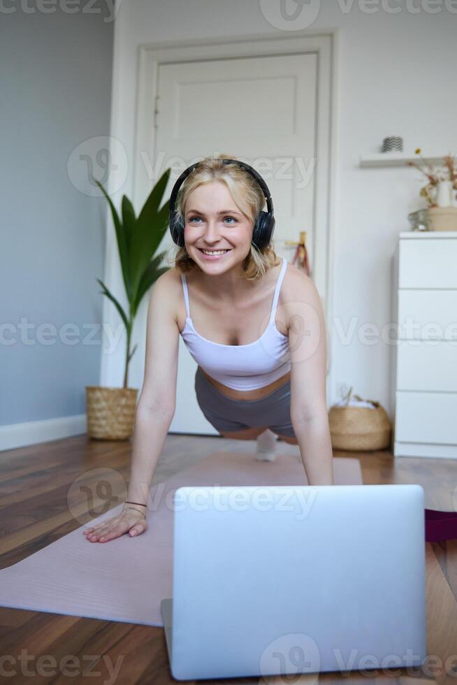 Portrait of young athletic woman in wireless headphones, standing in plank, following online workout , using laptop to join training session on the internet photo