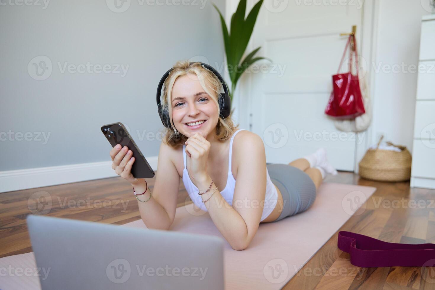 Portrait of young woman workout, watching exercise s on laptop in headphones, lying on rubber mat with mobile phone and smiling photo