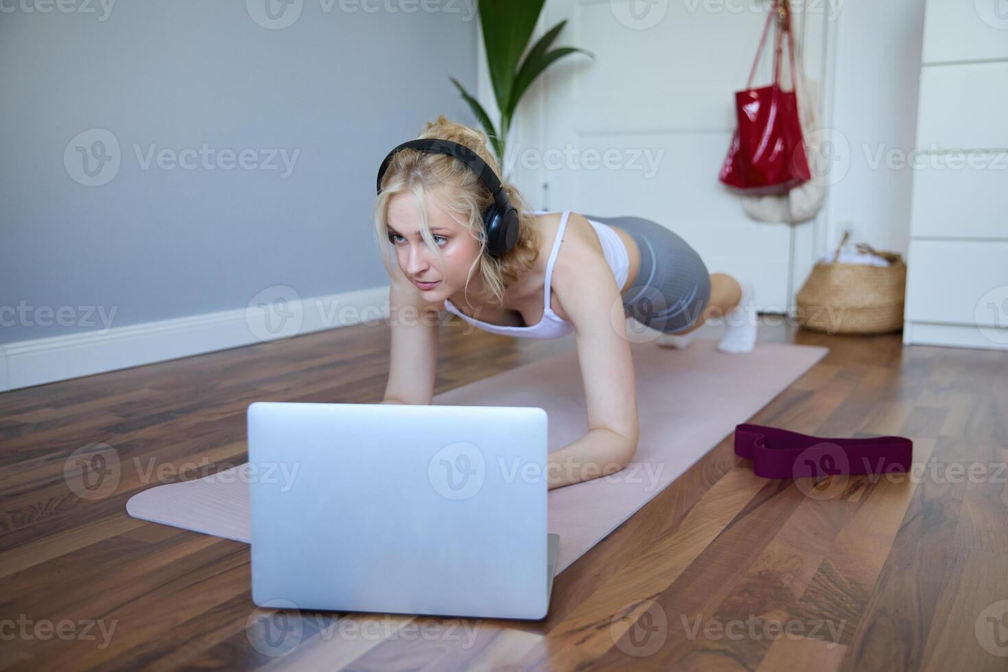 Portrait of young sporty woman doing workout, looking at fitness on laptop in wireless headphones, standing in plank on yoga mat, following exercise tutorial photo