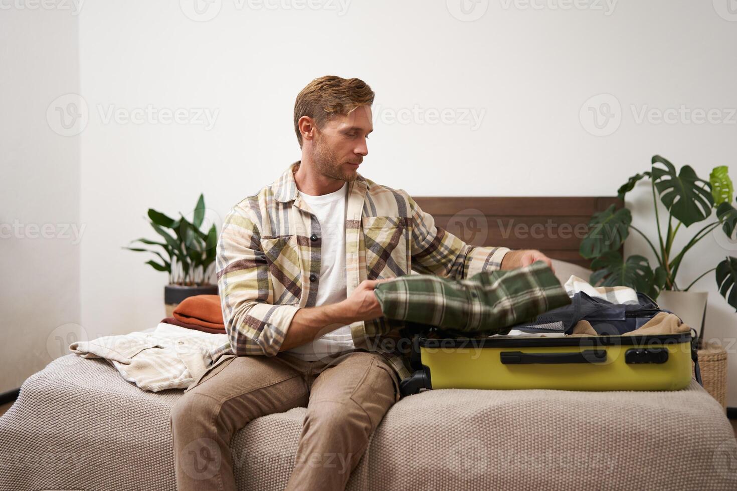 Portrait of young man packing his luggage for a trip, going on holiday, sitting on bed and putting clothes in suitcase, preparing for his flight photo