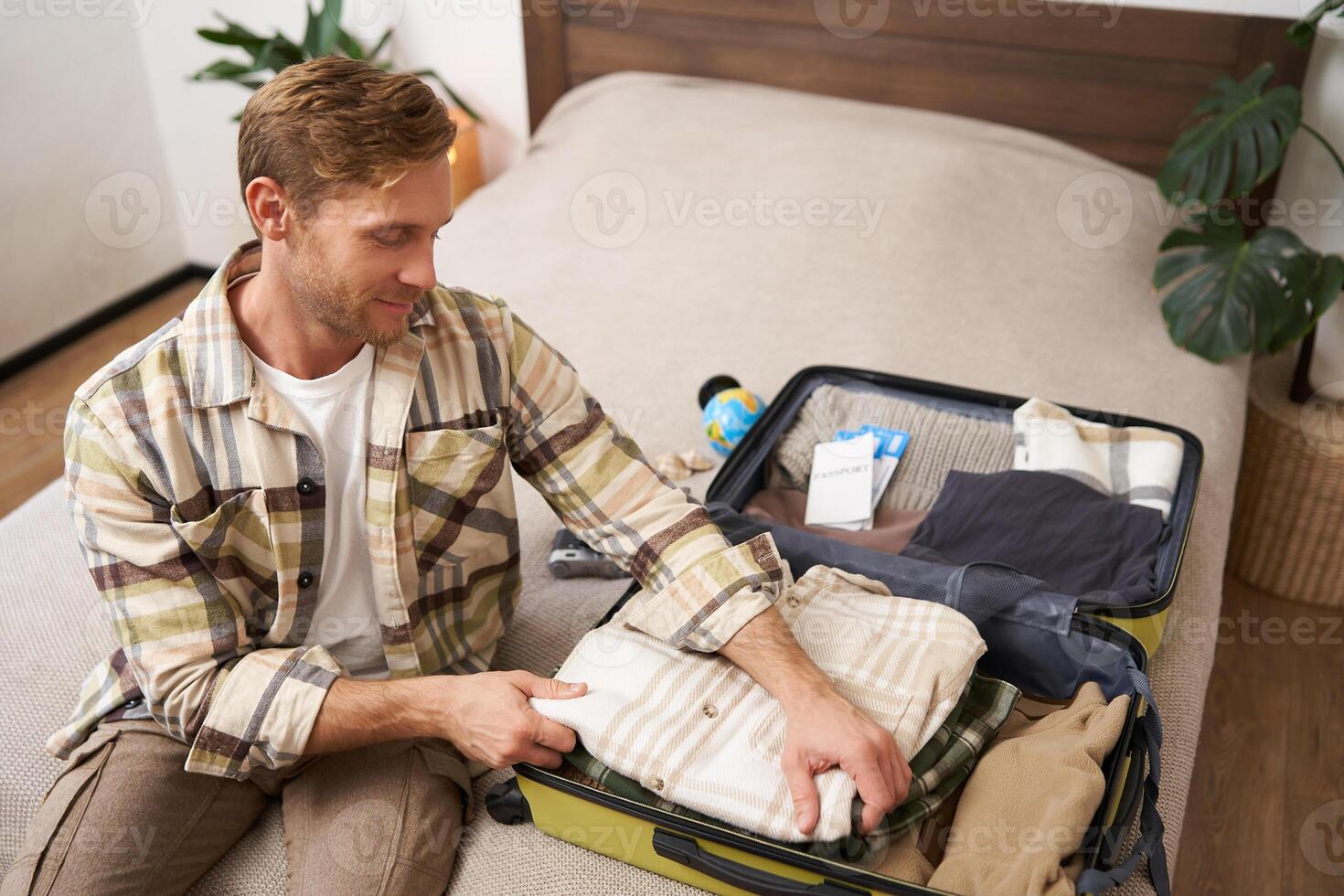 Portrait of handsome young man going on holiday, guy prepares for a trip journey, packs his suitcase, puts clothes in luggage, gets ready for adventure photo