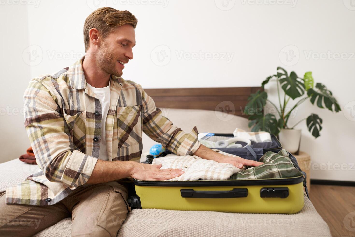 Portrait of handsome young man going on holiday, guy prepares for a trip journey, packs his suitcase, puts clothes in luggage, gets ready for adventure photo