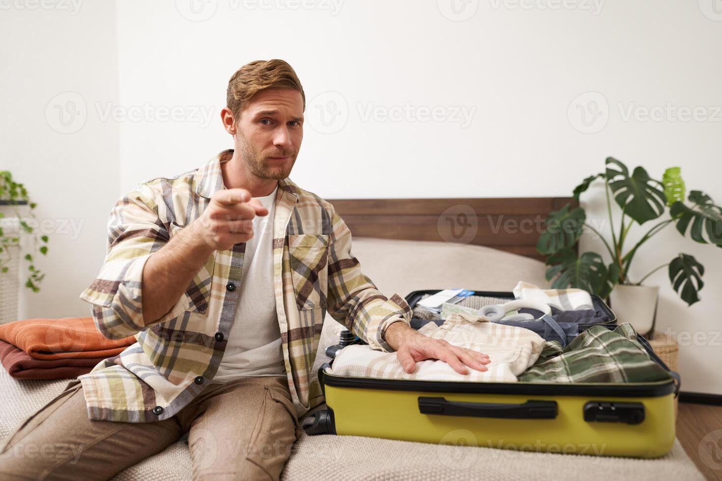 Portrait of handsome young man pointing finger at you, showing his packed suitcase, getting ready to go on holiday, encourages to contact travel agency and prepare for vacation photo