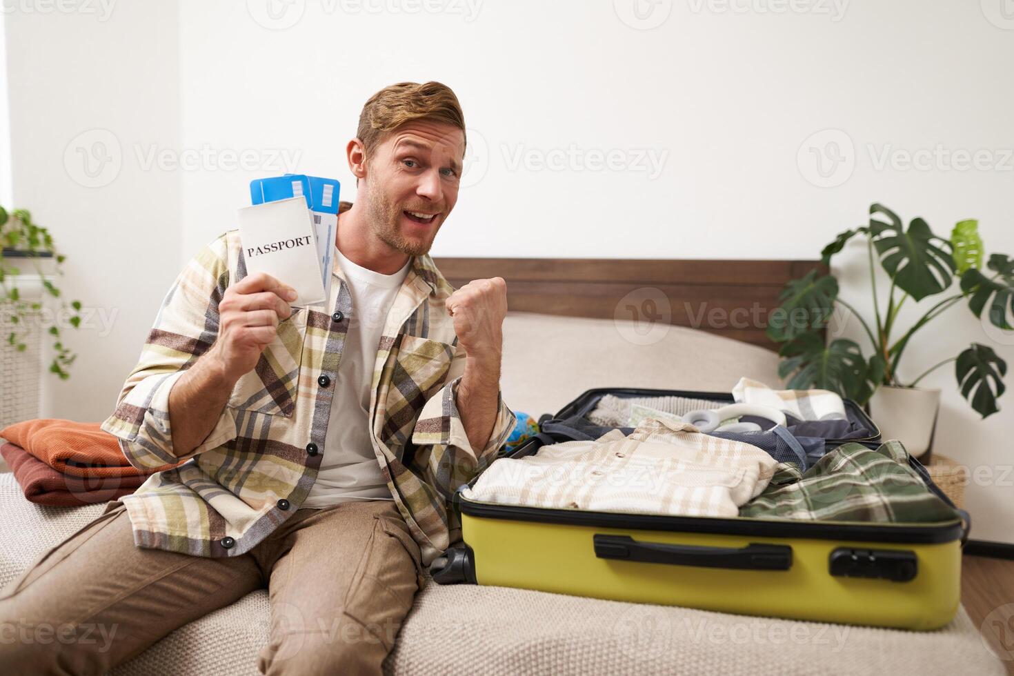 Portrait of cheerful guy sits near his luggage, packs suitcase, holds two plane tickets and passport, goes on holiday, books vacation stay photo